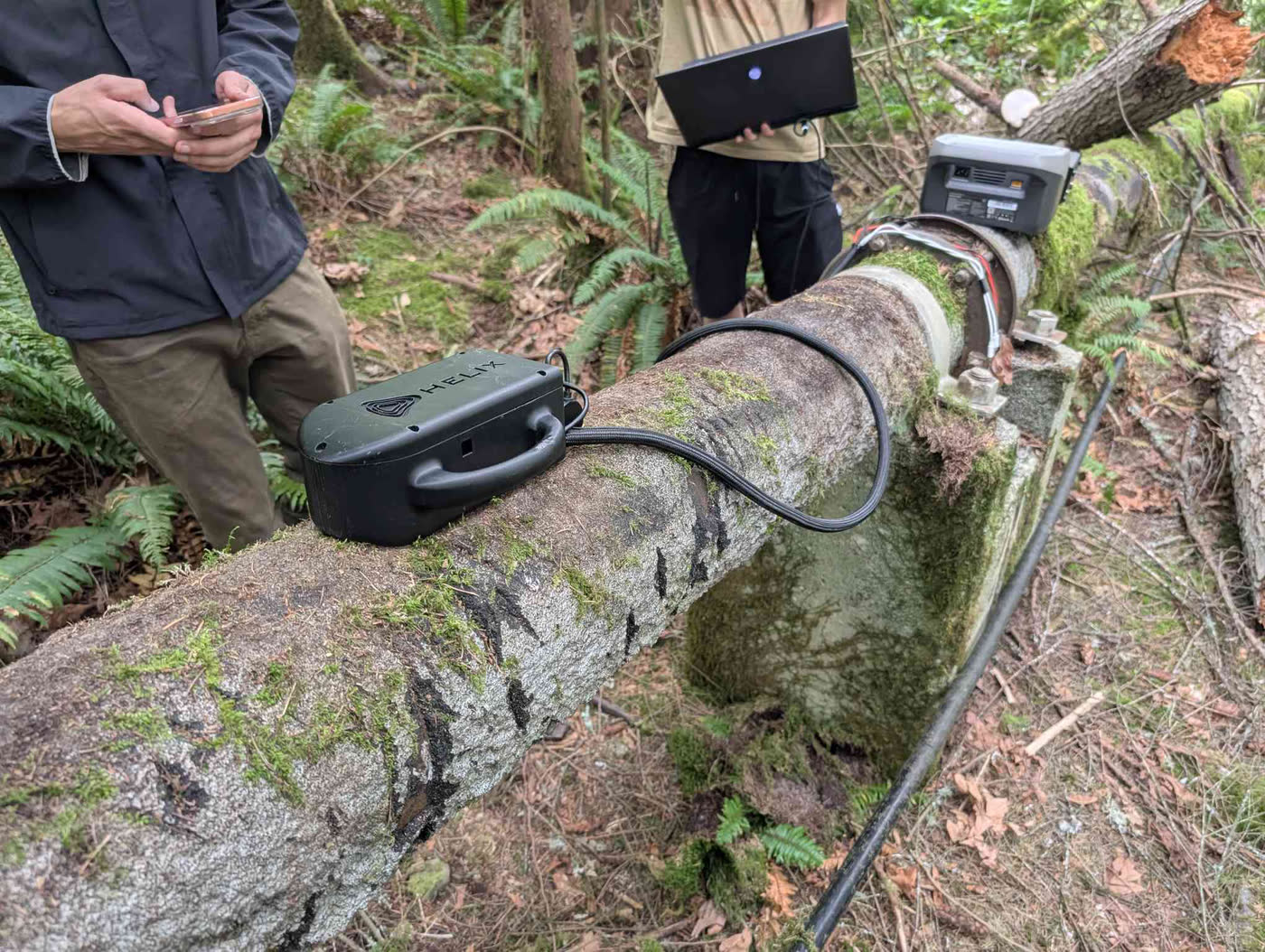 Two HELIX crew members screening a remote outdoor line with handheld equipment, the line still in service